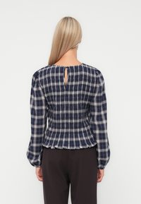 Donna bionda che indossa una camicia a maniche lunghe a quadri blu navy e bianca, con vita arricciata e chiusura a bottoni a forma di chiave sulla schiena.