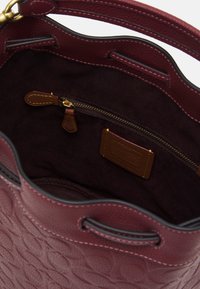 La borsa a secchiello in pelle texturizzata bordeaux presenta un interno con fodera in suede scuro, una tasca con cerniera e rifiniture in metallo dorato.