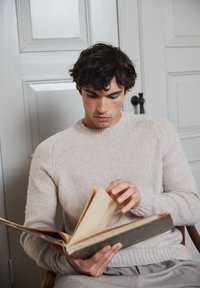 Suéter de punto beige claro con puños acanalados, llevado por una persona sentada, leyendo un gran libro abierto con páginas amarillentas.