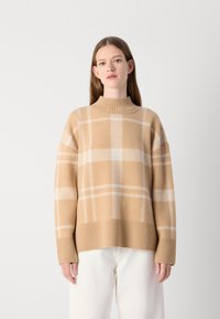 Suéter de malha bege com um decote alto, apresentando um padrão quadriculado branco. Punhos e barra canelados acrescentam textura. Combinado com calças brancas.