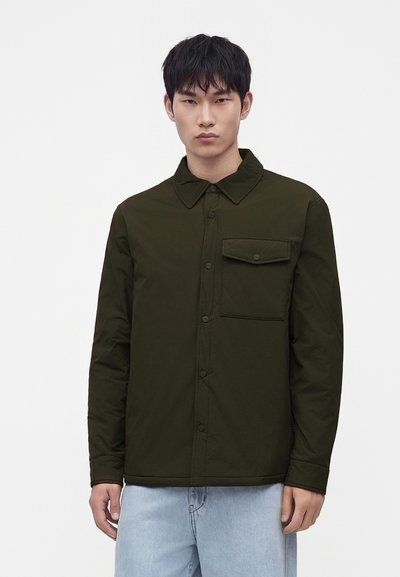 Jeune homme portant une veste boutonnée vert foncé avec une seule poche poitrine et un jean bleu clair, debout devant un fond uni.