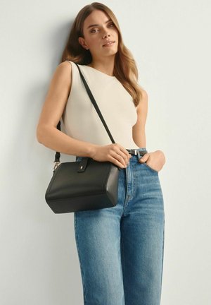 Kobieta ubrana w beżowy, bezrękawnik i niebieskie dżinsy, trzymająca czarną skórzaną torbę na ramię, z jedną ręką w kieszeni dżinsów.