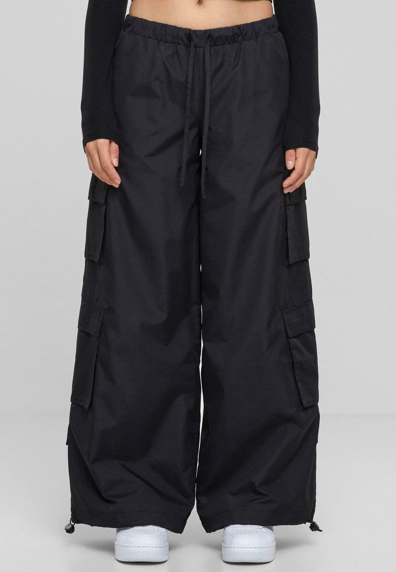 Pantalon cargo noir à jambes larges avec plusieurs poches, taille élastique avec cordon, porté avec des baskets blanches et un haut noir à manches longues.