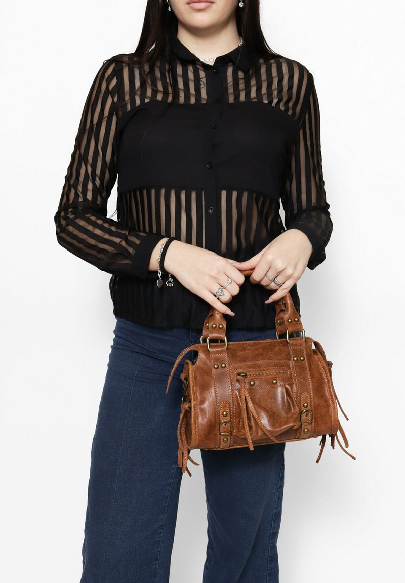 Piccola borsa a tracolla in pelle marrone con due manici, borchie decorative e dettagli frangiati. Indossata con una blusa trasparente nera a righe e jeans in denim.