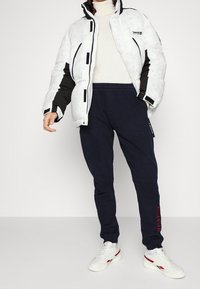 Puffy white and black jacket with a high collar, paired with dark navy sweatpants and white sneakers with red accents.
