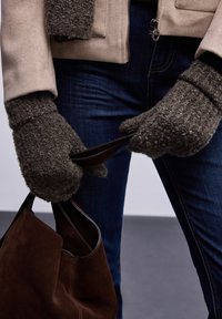 Moufles en polaire marron avec un détail en tricot texturé, portées en tenant un sac à main en daim marron. L'arrière-plan présente un sol gris foncé. Denim visible.