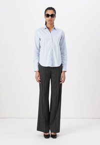 Camicia a righe azzurra chiara con bottoni, pantaloni a righe bianchi e neri a gamba larga e scarpe nere. Il modello indossa occhiali da sole scuri.