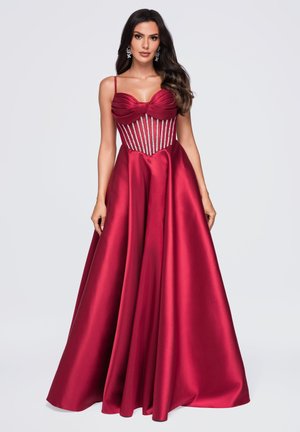 Femme aux longs cheveux foncés portant une robe en satin rouge longue jusqu’au sol avec un corsage style corset orné de perles et de fines bretelles.