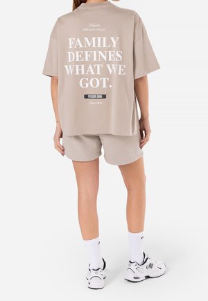 Person iført beige stor t-shirt med teksten "FAMILY DEFINES WHAT WE GOT", matchende shorts, hvide sokker og hvide sneakers, stående.