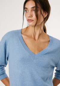 Šviesiai mėlynas v-kaklo megztinis, pagamintas iš minkštos mezginio medžiagos, su glotniais rankogaliais ir apačia, laisvo silueto bei lygiu paviršiumi.