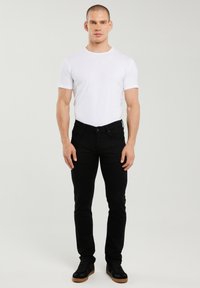 Mann steht frontal, trägt ein eng anliegendes weißes T-Shirt, schwarze schmale Jeans und schwarze Sneaker mit Gummisohlen, neutraler Hintergrund.