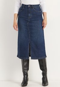 Jupe midi en denim bleu foncé avec une fente avant, cinq poches et des surpiqûres contrastées. Associée à des bottes noires jusqu'aux genoux.