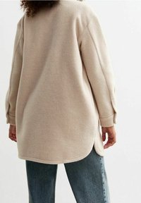 Sudadera de felpa beige de corte holgado con dobladillo curvado y hombros caídos, combinada con jeans de mezclilla azules, vista desde la parte de atrás.