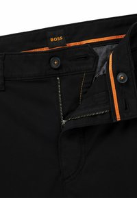 Pantaloni negri cu fermoar și închidere cu nasture. Cusături și căptușeală notabile de culoare portocalie, cu un etichetă din material care afișează "BOSS."