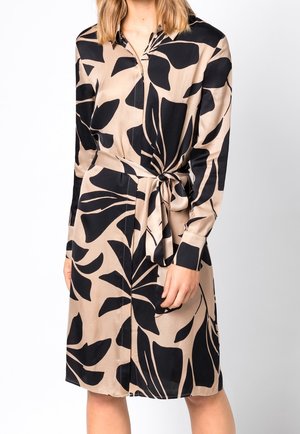 Femme portant une robe-chemise beige avec de grands motifs abstraits de feuilles noires et une ceinture nouée à la taille, debout devant un fond blanc.
