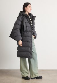Abrigo tipo puffer negro con capucha, con acolchado horizontal, botones frontales y un corte relajado, combinado con jeans de pierna ancha en verde claro y zapatos de plataforma.