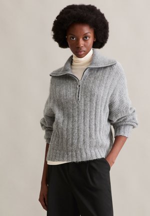 Grauer gerippter Strickpullover mit hohem Kragen, Viertelreißverschluss vorne und aufgerollten Ärmeln. Über einem cremefarbenen Rollkragenpullover getragen.