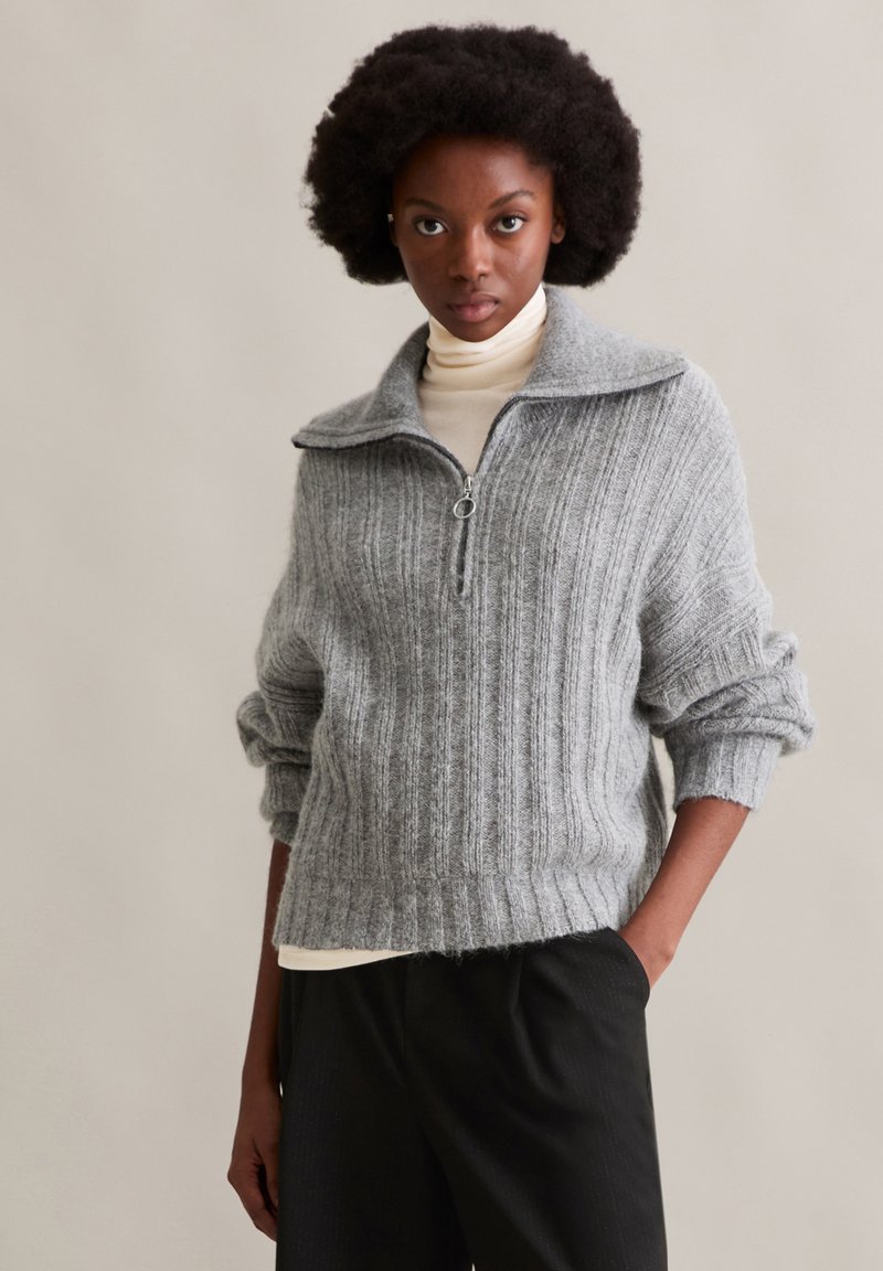 Szary sweter dzianinowy z prążkami, z wysokim kołnierzem, zamek błyskawiczny na ćwierć przodu i podwiniętymi rękawami. Nałożony na kremowy golf.