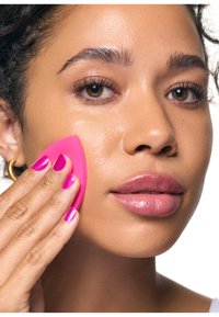 Kobieta z brązowymi oczami nakłada makijaż różową gąbką na policzek, prezentując gładką skórę i błyszczące usta.