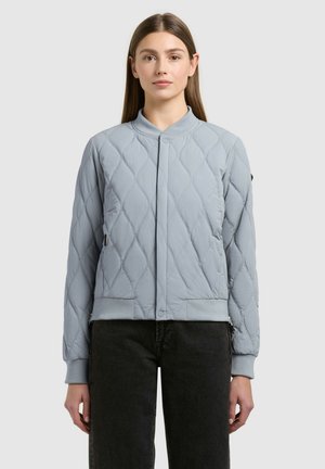 Woman wearing a light blue quilted bomber jacket with snap button closure and black pants, standing against a plain gray background.