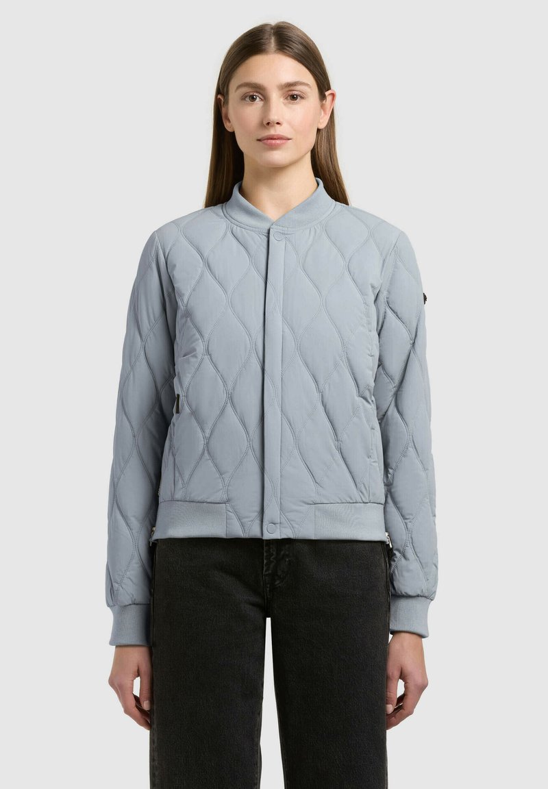 Femme portant une veste bomber matelassée bleu clair avec fermeture à boutons-pression et un pantalon noir, debout devant un fond gris uni.