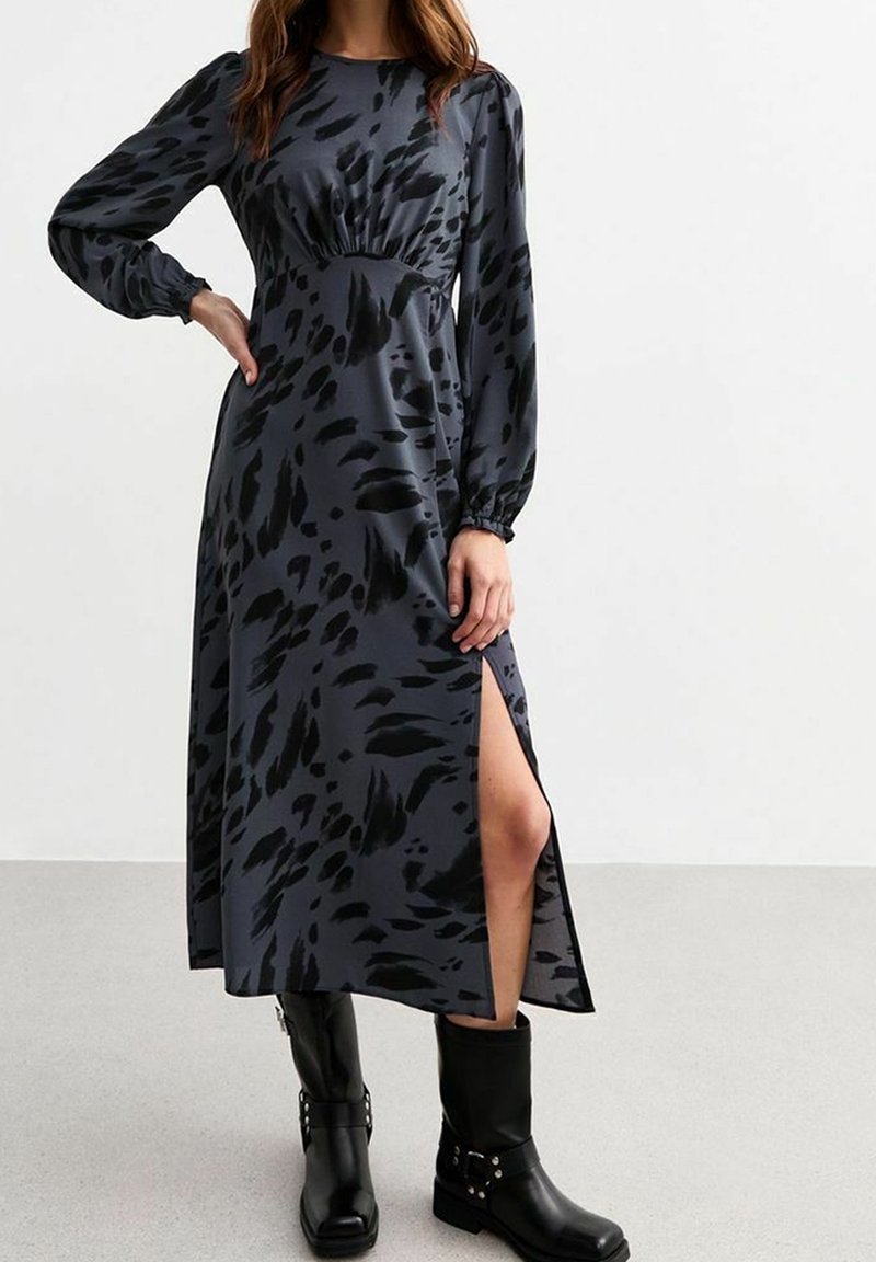 Femme portant une longue robe gris foncé avec des taches abstraites noires, manches longues, fente latérale et bottes noires mi-mollet, debout à l'intérieur.