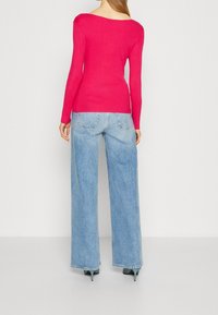 Eine Frau, die von hinten steht, trägt ein leuchtend pinkes Langarmoberteil, helle, weit geschnittene Jeans und türkisfarbene High Heels und lehnt an einem schlichten Hintergrund.