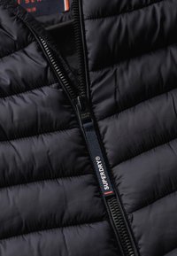 Sort pufferjakke med lynlås, quiltet tekstur og et mærke med brandnavnet "Superdry." Glat stoffinish.