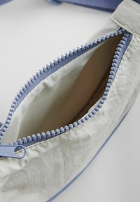 Borsa grigia chiara con zip blu e nastro di trazione, caratterizzata da una superficie texture e una fodera interna a contrasto. L'apertura con zip è visibile.