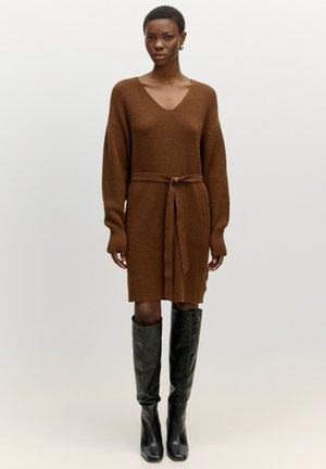 Bruine gebreide jurk met V-hals, lange mouwen en een afneembare strikriem. Gestyled met zwarte leren laarzen tot de knie.