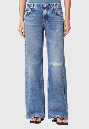 Blå jeans med vide ben, høj talje og let slidt plet på højre lår, båret med sorte sandaler.