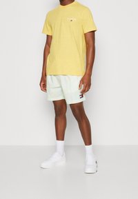 Uomo che indossa una maglietta gialla di Tommy Jeans, pantaloni corti camo chiari in tonalità pastello con logo, calzini bianchi e sneakers bianche, in piedi contro uno sfondo semplice.