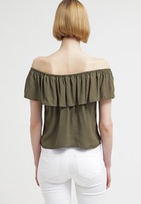 Olivenfarvet off-the-shoulder top med flæse i halsen og løs pasform. Lavet af letvægtsstof. Bæres med hvide tætsiddende bukser.