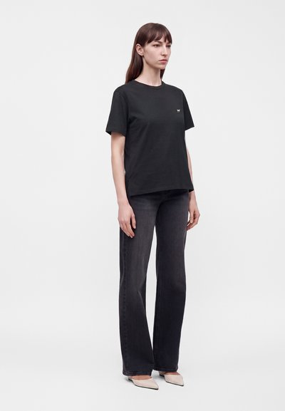 Jeune femme debout portant un t-shirt noir uni à manches courtes, un jean large foncé et des chaussures pointues de couleur claire, sur un fond uni.