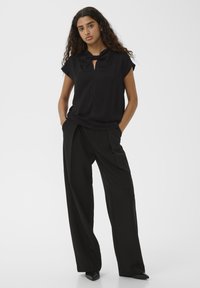 Donna con lunghi capelli ricci che indossa una maglietta nera a maniche corte con dettaglio al collo a fiocco e pantaloni neri a gamba larga, mani nelle tasche.