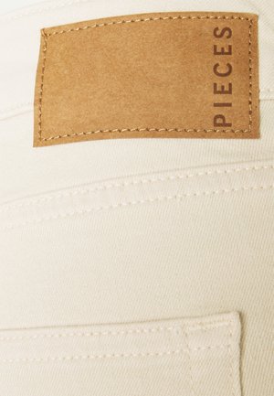 Beige stof met een getextureerde afwerking, voorzien van een bruine leren label met het woord "PIECES" erop gestikt. Zichtbare stiksels en een glad ontwerp.