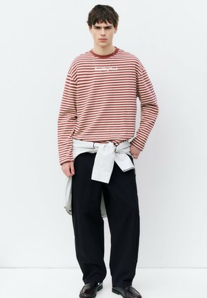 Jeune homme avec une chemise marron à rayures manches longues, pantalon noir ample, chemise blanche nouée autour de la taille, debout devant un fond blanc uni.