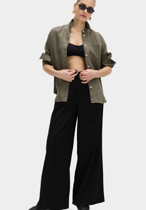 Vrouw draagt zwarte wijde broek, zwart bandeautopje, olijfgroen open overhemd, zonnebril en gouden oorringen, en stelt de taille van haar broek bij.