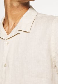 Camicia di lino beige chiaro con colletto corto e morbido, con chiusura a bottoni frontale, realizzata in un tessuto testurizzato e leggermente stropicciato.