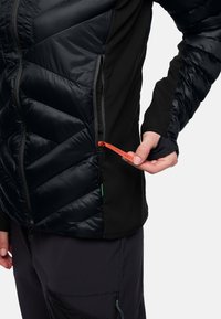 Persona che indossa una giacca trapuntata nera mentre apre una tasca laterale con un tirazip rosso etichettato "PACK AWAY."