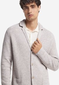 Jasnoszary kardigan dziany z kołnierzem szalowym, fakturowany materiał, trzy guziki i dwie przednie kieszenie, noszony na kremowej koszuli.