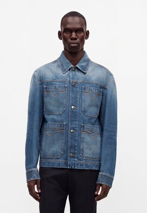 Mand iført en blå denimjakke med fire forlommer og sølvknapper, kombineret med sorte bukser, stående mod en hvid baggrund.