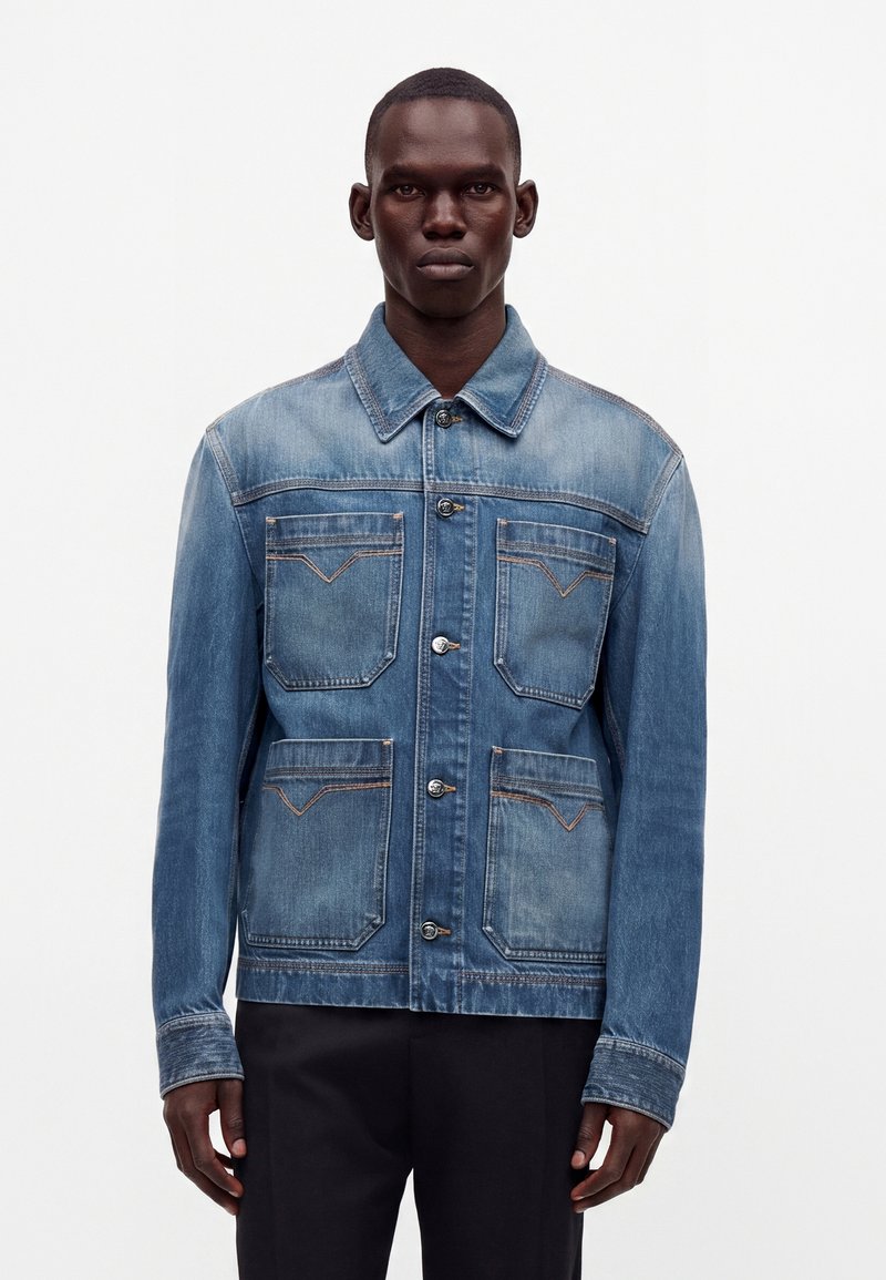 Homme portant une veste en jean bleue avec quatre poches avant et des boutons argentés, associée à un pantalon noir, debout devant un fond blanc.