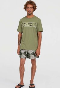 Tamsiai žalia marškinė su grafiniu spaudiniu, derinami su raštuotomis šortais žaliai ir baltai gėlėta dizainu. Nešiojami su juodais sandalais.