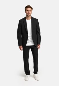 Blazer negro a medida con solapas de muesca, dos bolsillos frontales, combinado con una camiseta blanca y pantalones negros. Zapatillas blancas completan el look.