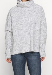 Grau turtleneck Pullover mit lockerer Passform, gerippter Textur und fehlenden Schultern. Er hat einen rohen Saum und passt gut zu blauen Jeans.