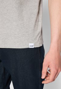 Pelēka kokvilnas t-krekla ar apaļu apkakli un īsām piedurknēm, uz kuras apakšmalas ir maza balta "Wrangler" birka, pārī ar tumšiem džinsiem.