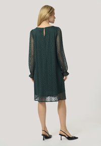Green lace dress with long, sheer sleeves, diamond pattern, and back keyhole detail, paired with black slingback heels.