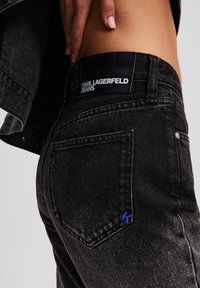Vaqueros de denim negro con un acabado texturizado, que cuentan con un parche de logo negro en la cintura y un pequeño acento azul 'K' en el bolsillo trasero.