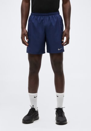 Uomo che indossa pantaloncini Nike blu navy, scarpe da ginnastica nere e calzini bianchi Nike, in piedi davanti a uno sfondo bianco.